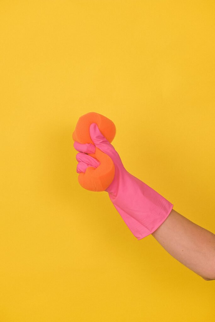 A pink gloved hand squeezes an orange sponge on a bright yellow background, emphasizing cleanliness and hygiene.